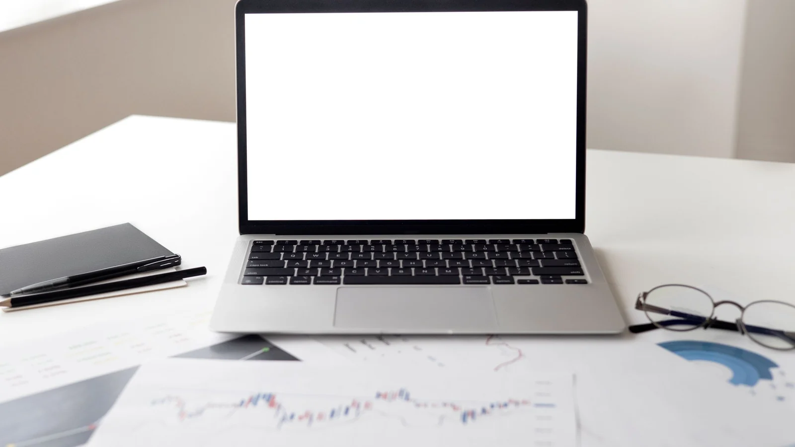 Notebook aberto entre relatórios, caderno, canetas e óculos sobre uma mesa de trabalho.