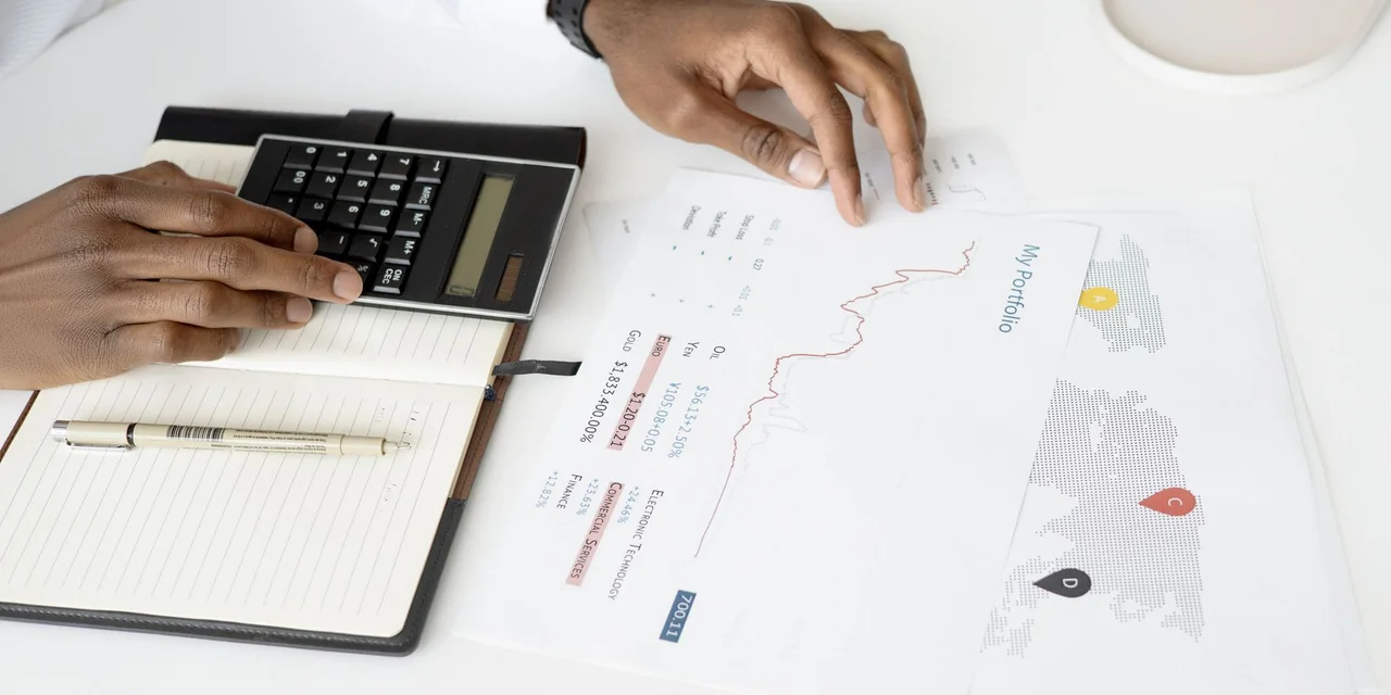 Mãos usando uma calculadora ao lado de caderno e relatórios sobre uma mesa branca.